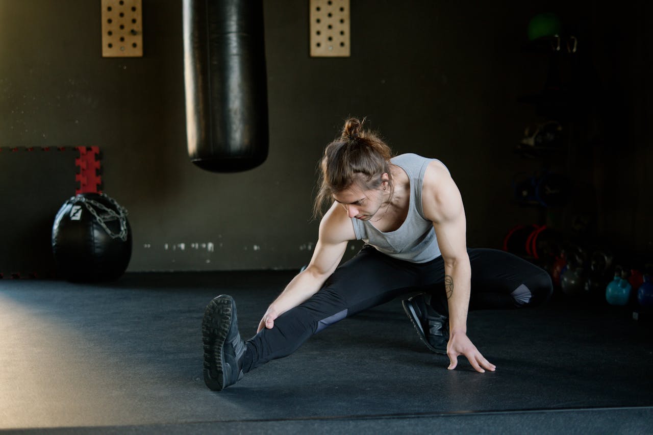 A fit young man stretches in a gym with a punching bag and fitness equipment in the background.