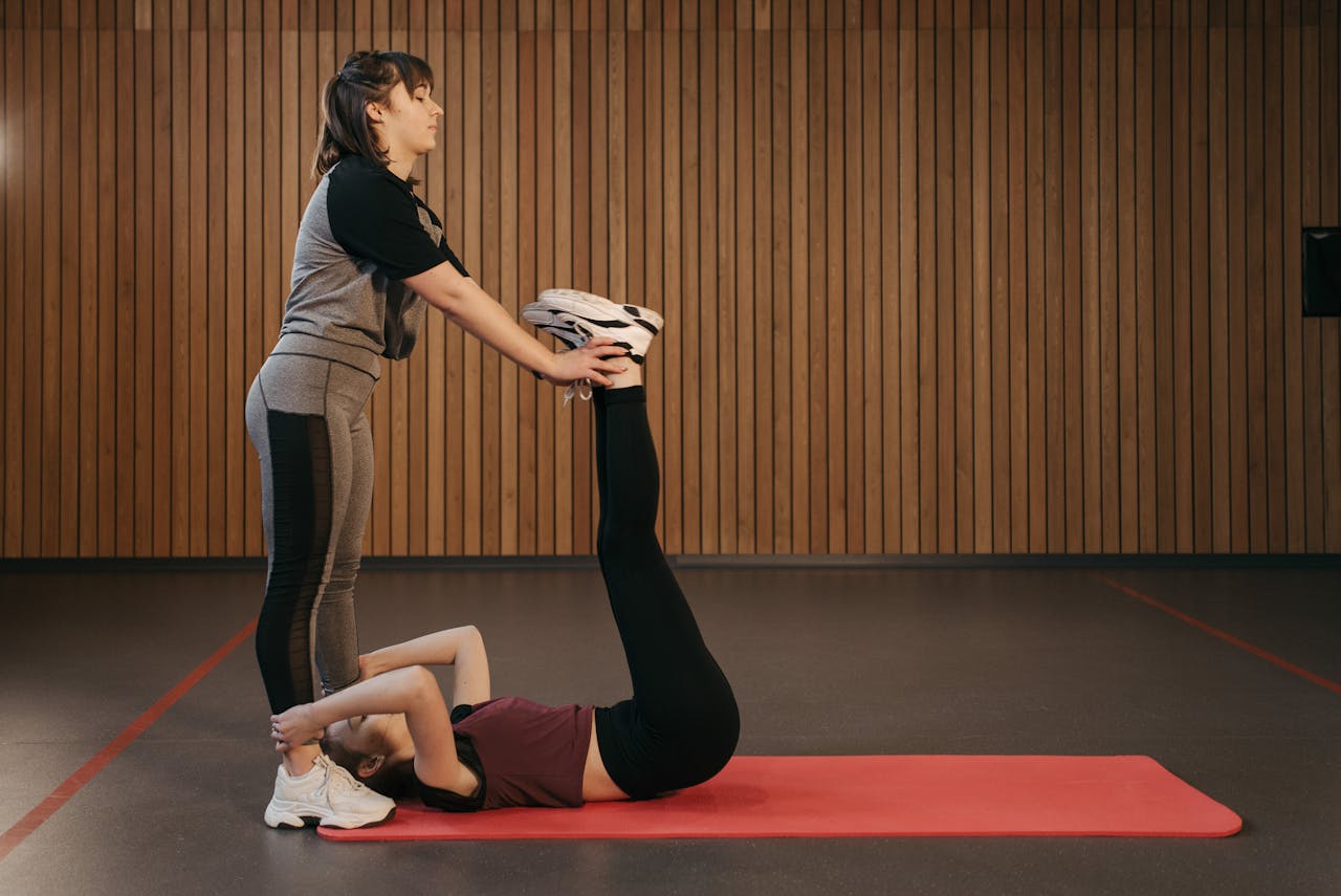 Two women engage in partner-assisted stretching on a mat in a gym setting, enhancing flexibility.